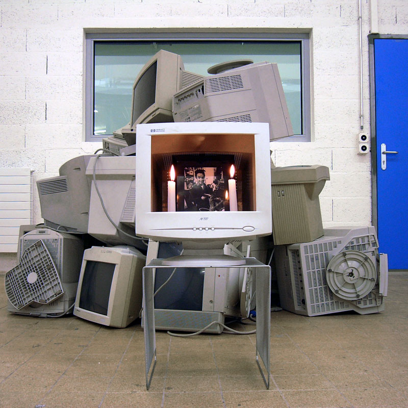 Photo of an empty CRT screen with two candles and a picture of Nam June Paik inside, in front of a pile of CRT screens